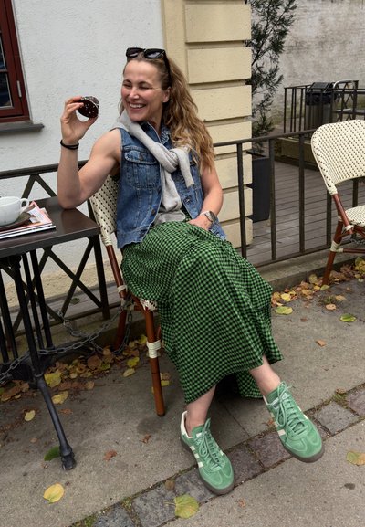 Chaleco de mezclilla sobre sudadera gris, falda a cuadros verdes y zapatillas verdes. Sosteniendo un donut de chocolate, sentado en una mesa con una taza.