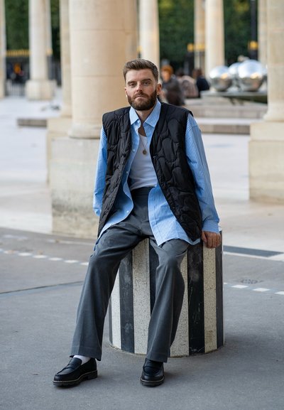 Chaleco acolchado negro sobre una camisa a rayas azul y blanca, pantalones grises y mocasines negros, sentado sobre una columna de piedra a rayas al aire libre.