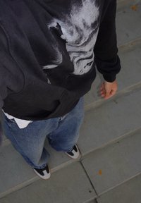 Melnas kapuces jaciņa ar pelēku abstraktu rakstu, baltu T-kreklu kā apakšējo slāni, brīvi pieguļošas zilas džinsa bikses un melni balti sporta apavi.