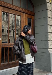 Brun skinnjakke i stor størrelse, grønn strikkegenser, hvit bluse med rysjer, svart skjørt med polkaprikker og en burgunder veske. Hodetelefoner båret på hodet.