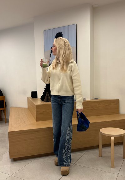 Sudadera blanca de cremallera con canalé, jeans azules con bordado floral, zuecos beige de plataforma, bolso azul, sosteniendo un vaso con una bebida verde.
