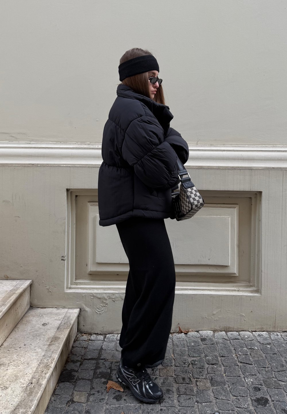 Giubbotto imbottito nero con vita aderente, indossato sopra pantaloni neri a gamba larga. Completato da una borsa con motivo e occhiali da sole scuri.