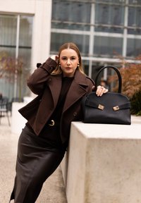 Ruskea nahkalaukku, jossa on kultaiset metalliosat, kestävä kahva ja jämäkkä muotoilu; sijoitettu vaalealle kivipinnalle.