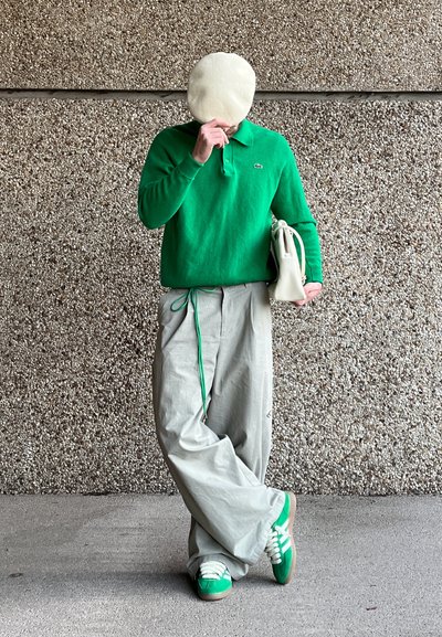 Suéter verde texturizado, pantalones grises sueltos, acentos verdes, zapatos casuales con cordones blancos, sosteniendo una bolsa blanca. El fondo es una superficie texturizada.