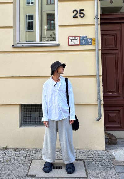 Camisa blanca de botonadura, pantalones de chándal grises, gorra de cubo negra y chanclas negras de peluche, con una pared beige texturizada y características de una puerta de madera.