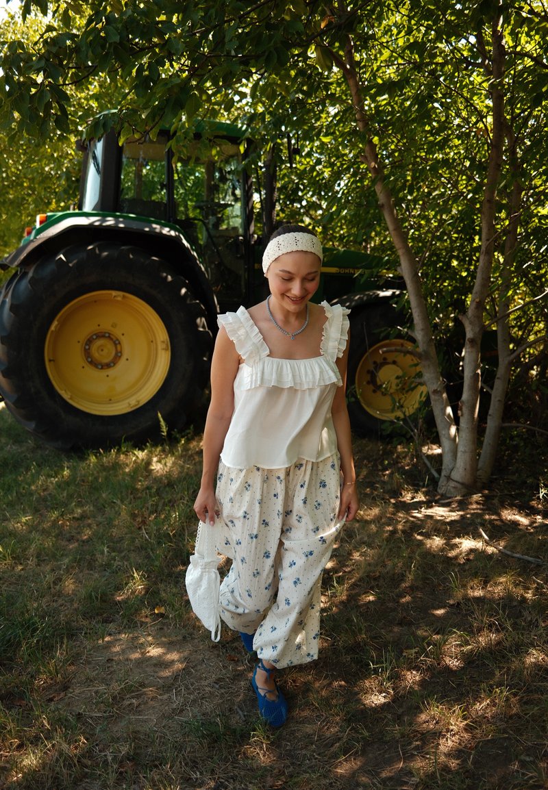 Jeune femme portant un pantalon fleuri et un haut à volants marche à l’extérieur près d’un tracteur vert sous des arbres feuillus, tenant un sac blanc.
