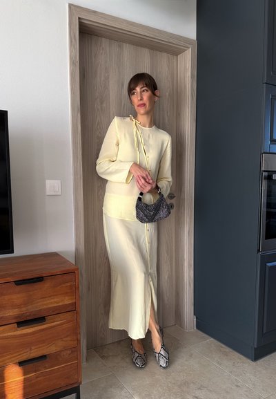 Blusa amarilla abotonada con detalle de lazo, combinada con una falda midi a juego. Bolso de mano con estampado en blanco y negro, zapatos de piel de serpiente. Ambiente interior moderno.