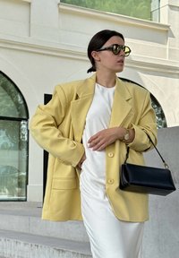 Dzeltena pārāk liela bleizera ar lieliem pogām, uzvilkta virs baltas kleitas. Melna somiņa piešķir kontrastu. Modelis valkā tumšus saulesbrilles.