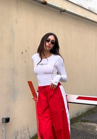 Jeune femme portant des lunettes de soleil, un haut à manches longues blanc et un pantalon de survêtement rouge, appuyée contre une barrière dans un environnement urbain en plein air.