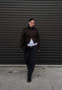 Brun ribbestrikket genser med høy krage, kombinert med løse svarte jeans. Holder en svart veske, står mot en mørk teksturert vegg.