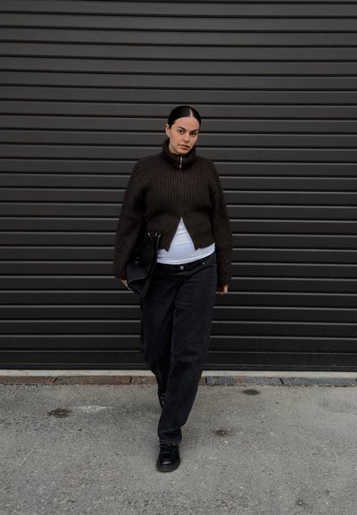 Suéter marrón de punto acanalado con cuello alto, combinado con jeans negros holgados. Sosteniendo una bolsa negra, de pie frente a una pared oscura con textura.