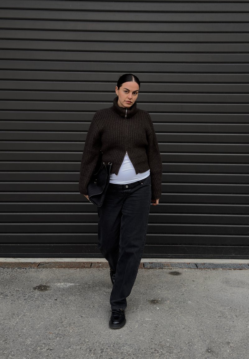 Brauner gerippter Strickpullover mit hohem Kragen, kombiniert mit lockeren schwarzen Jeans. Hält eine schwarze Tasche und steht vor einer dunklen, strukturierten Wand.