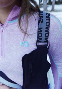 Lys lilla genser med glidelås, med subtilt mønster, blå aksenter og en svart skulderrem med teksten "WOMOW." Svart lomme med glidelåsdetalj.