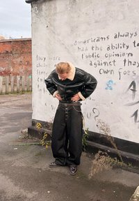 Personne portant une veste en cuir noire et un pantalon noir surdimensionné, debout à l'extérieur près d'un mur blanc couvert de graffitis avec un texte pâle.