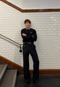 Polo cu mânecă lungă de culoare navy cu guler bleu deschis, curea gri, pantaloni largi negri, pantofi negri. Pe fundalul unui zid de faianță albă pentru metrou și scări.