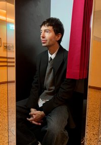 Osoba koja sjedi u crnom odijelu i sivoj dolčeviti ispred crne pozadine, s crvenom zavjesom s jedne strane i sivoj podnoj pločici.