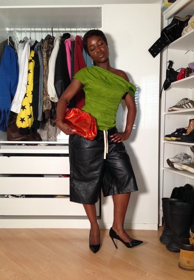 Top verde sin hombros con pliegues, culottes de cuero negro con cordón, billetera naranja y tacones negros de punta sobre un fondo de ropa.