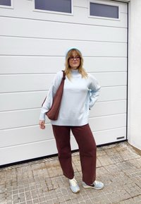 Femme portant un pull bleu clair, un bonnet bleu, un pantalon marron, des baskets et des lunettes, se tenant avec un sac marron sur l'épaule devant une porte de garage blanche.