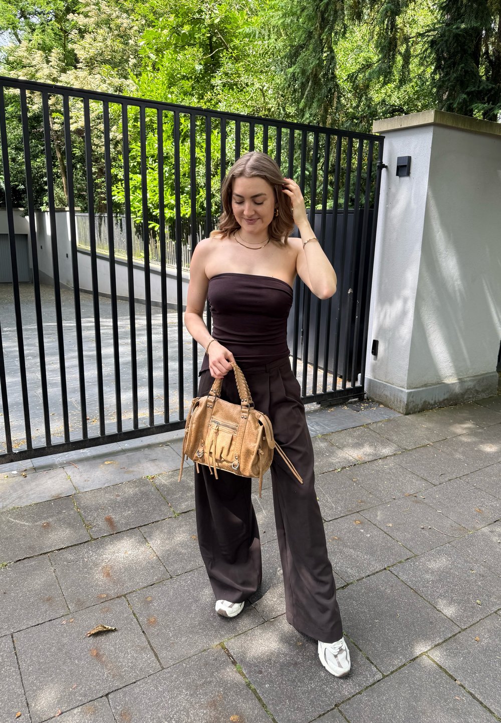 Svart strapless topp, mörka vida byxor och vita sneakers. Håller en beige handväska med flätad handtag och dragkedjedetalj.