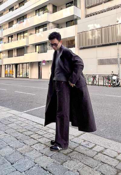 Abrigo largo negro sobre un suéter y pantalones oscuros, con sandalias negras. Entorno urbano, acera de adoquines, edificios modernos de fondo.