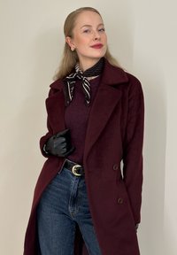 Manteau bordeaux à double boutonnage, gants noirs, haut violet foncé et jean en denim. Une écharpe en soie rayée noir et blanc souligne le look.