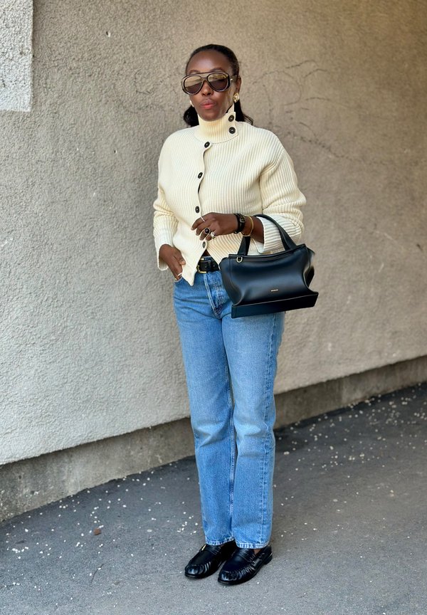 Kobieta w kremowym prążkowanym swetrze, niebieskich dżinsach, czarnych mokasynach, dużych okularach przeciwsłonecznych, trzymająca czarną torebkę, stojąca na tle teksturowanej ściany.
