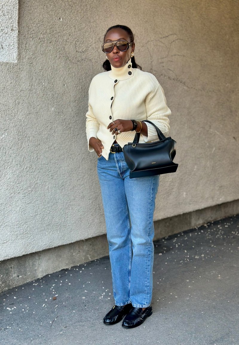 Frau trägt cremefarbenen Rippstrickpullover, blaue Jeans, schwarze Loafer, große Sonnenbrille, hält schwarze Handtasche, steht vor strukturierter Wand.