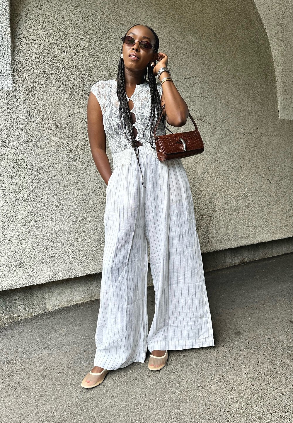 Λευκή, χωρίς μανίκια φόρμα με δαντελένια μπούστο, φαρδιά παντελόνια με λεπτές κάθετες ρίγες. Καφέ, υφαντή τσάντα και διαφανή παπούτσια.