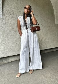 Λευκή, χωρίς μανίκια φόρμα με δαντελένια μπούστο, φαρδιά παντελόνια με λεπτές κάθετες ρίγες. Καφέ, υφαντή τσάντα και διαφανή παπούτσια.