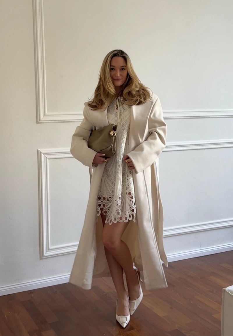 Frau mit langen Haaren steht drinnen, trägt ein weißes Spitzenkleid, einen beigen langen Mantel und weiße High Heels und hält eine beige Clutch-Tasche.