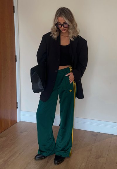 Blazer oversize negro sobre un top corto negro, combinado con pantalones deportivos de pierna ancha verdes con rayas amarillas. Bolso negro y botas de tobillo.