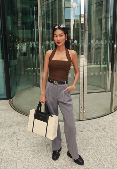 Mujer con top halter marrón, pantalones grises, cinturón negro y mocasines, sostiene un bolso beige y negro cerca de una puerta giratoria de cristal.