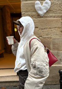 Personne portant une veste beige et un sweat à capuche blanc, tenant une boisson, avec un sac à main rouge à l'épaule, marchant devant un mur en pierre orné d'une décoration en forme de cœur blanc.