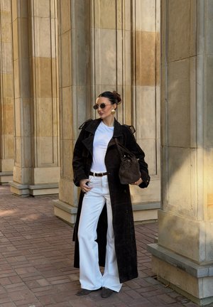 Schwarzer Wildleder-Trenchcoat, weißes T-Shirt, weiße weite Hose, runde Sonnenbrille, goldene Ohrringe, hält eine dunkle Handtasche, vor steinernen Säulen.