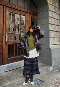 Brun skinnjakke over en grønn genser, hvit skjorte med rysjer og svart skjørt med hvite prikker. Hvite hæler og grå sokker fullfører antrekket.