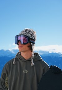 Lunettes de ski avec verres roses et monture noire. Bonnet tricoté gris avec rayures blanches et pompon. Sweat à capuche gris. Snowboard sous le bras.