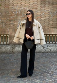 Jachetă bej din blană artificială cu bordură neagră, pulover maro închis cu nasturi, pantaloni negri cu picioare largi și o geantă mică neagră, pe fundalul unui zid de cărămidă.