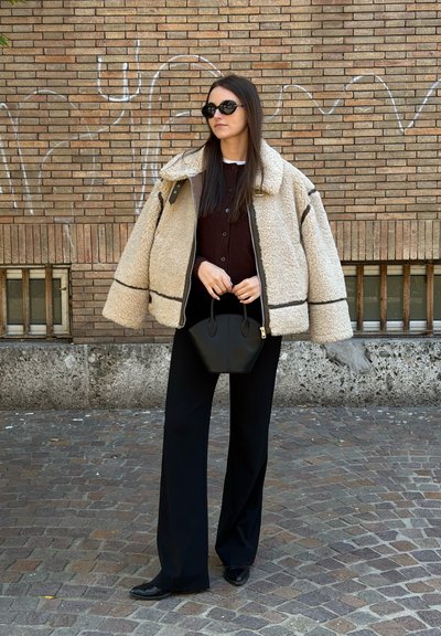 Chaqueta beige de borrego texturizado con ribetes oscuros, pantalones negros de pierna ancha y un pequeño bolso negro. Gafas de sol redondas negras completan el conjunto.