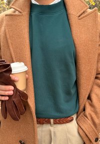 Manteau en laine marron sur un sweat-shirt bleu sarcelle, gants en cuir marron et ceinture beige. Tenant une tasse de café avec un couvercle.