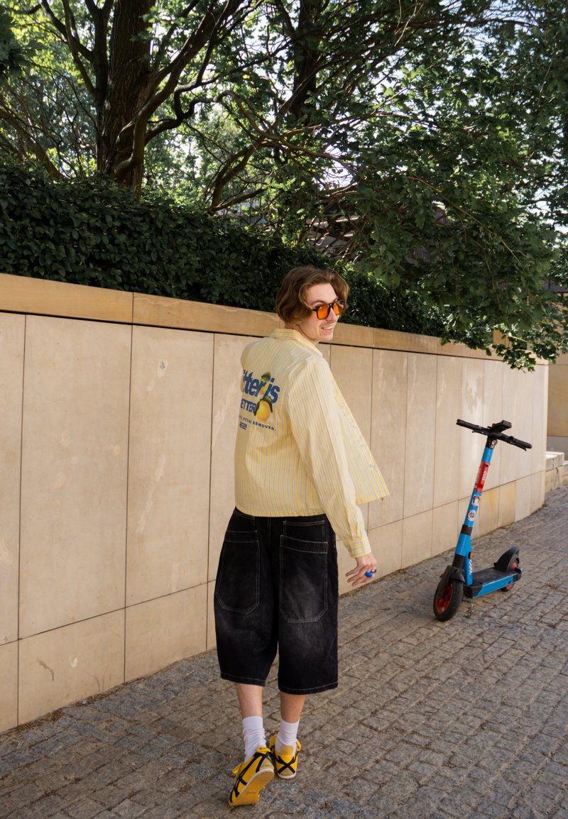 Personne en chemise rayée jaune et short noir marchant sur un chemin pavé près d'une trottinette électrique près d'un mur beige et d'un feuillage vert.
