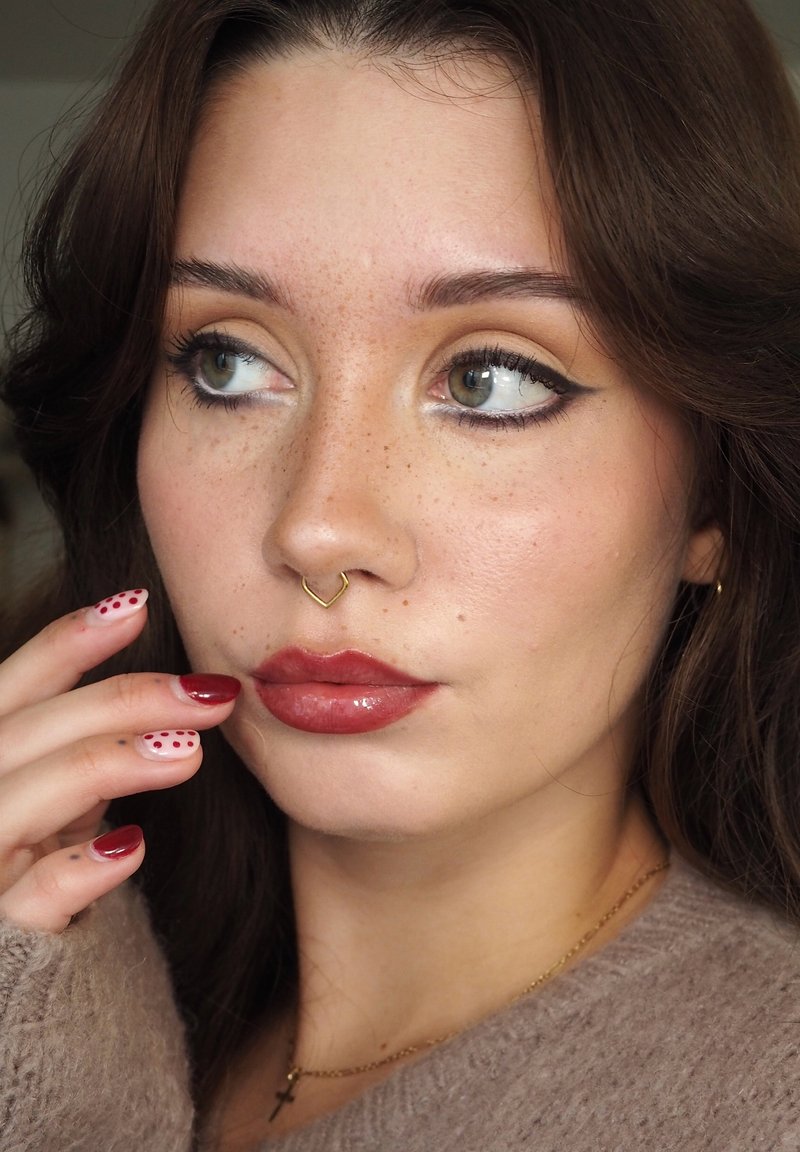Giovane donna con piercing al setto nasale, occhi verdi con eyeliner allungato, labbra rosse lucide e unghie dipinte di rosso e bianco con pois rossi.