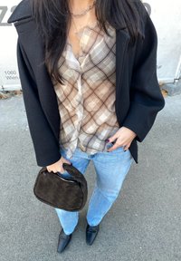 Mulher vestindo um blazer preto, blusa xadrez transparente, jeans azul claro e sapatos pretos apontados, segurando uma bolsa de palha preta numa rua.