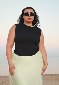 Top nero senza maniche con un design aderente abbinato a una gonna leggera verde chiaro. La modella indossa occhiali da sole scuri e ha i capelli ricci.