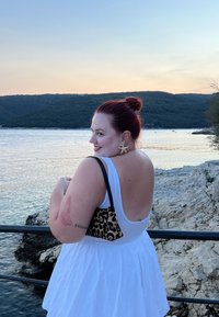 Frau in einem weißen ärmellosen Kleid mit offenem Rücken, schwarzer Tasche mit Leopardenmuster und sternförmigen Ohrringen, die bei Sonnenuntergang am Wasser steht.