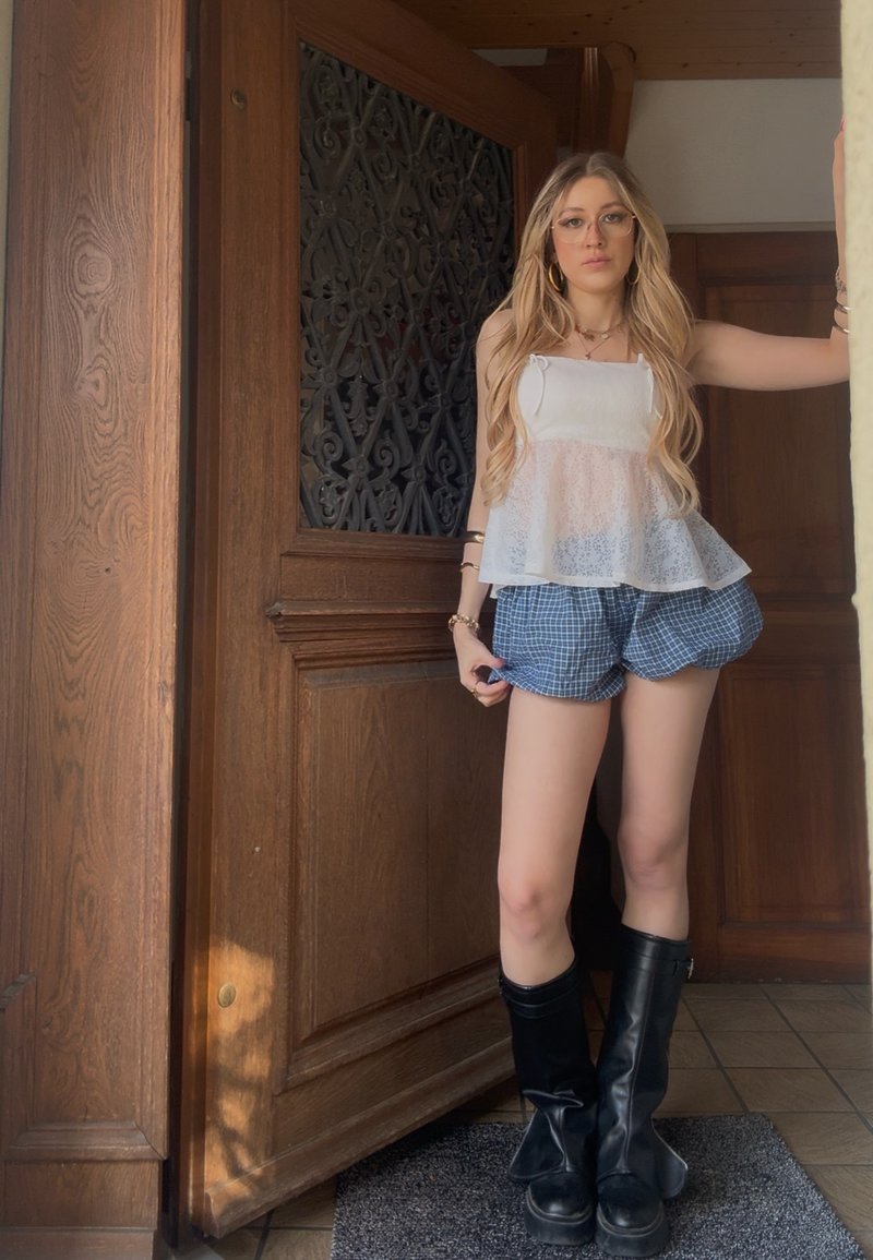 Blonde woman with long hair wearing glasses, a white sleeveless top, blue checkered shorts, and black boots, standing by a wooden door.