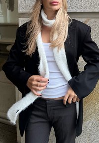Zwarte blazer, witte wollen sjaal en witte tanktop; grijze broek. Glad stof, warme textuur, gelaagde look met contrasterende kleuren.