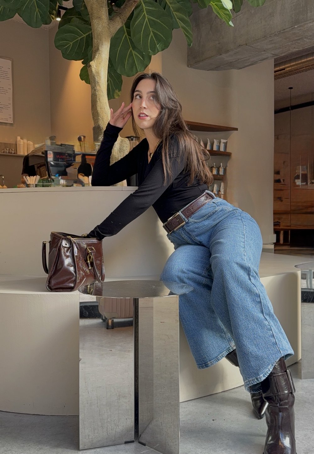 Maglietta nera a maniche lunghe, jeans larghi blu e stivali di pelle marrone. Borsa marrone con dettagli dorati. Tavolo bianco moderno e curvo con superficie riflettente.