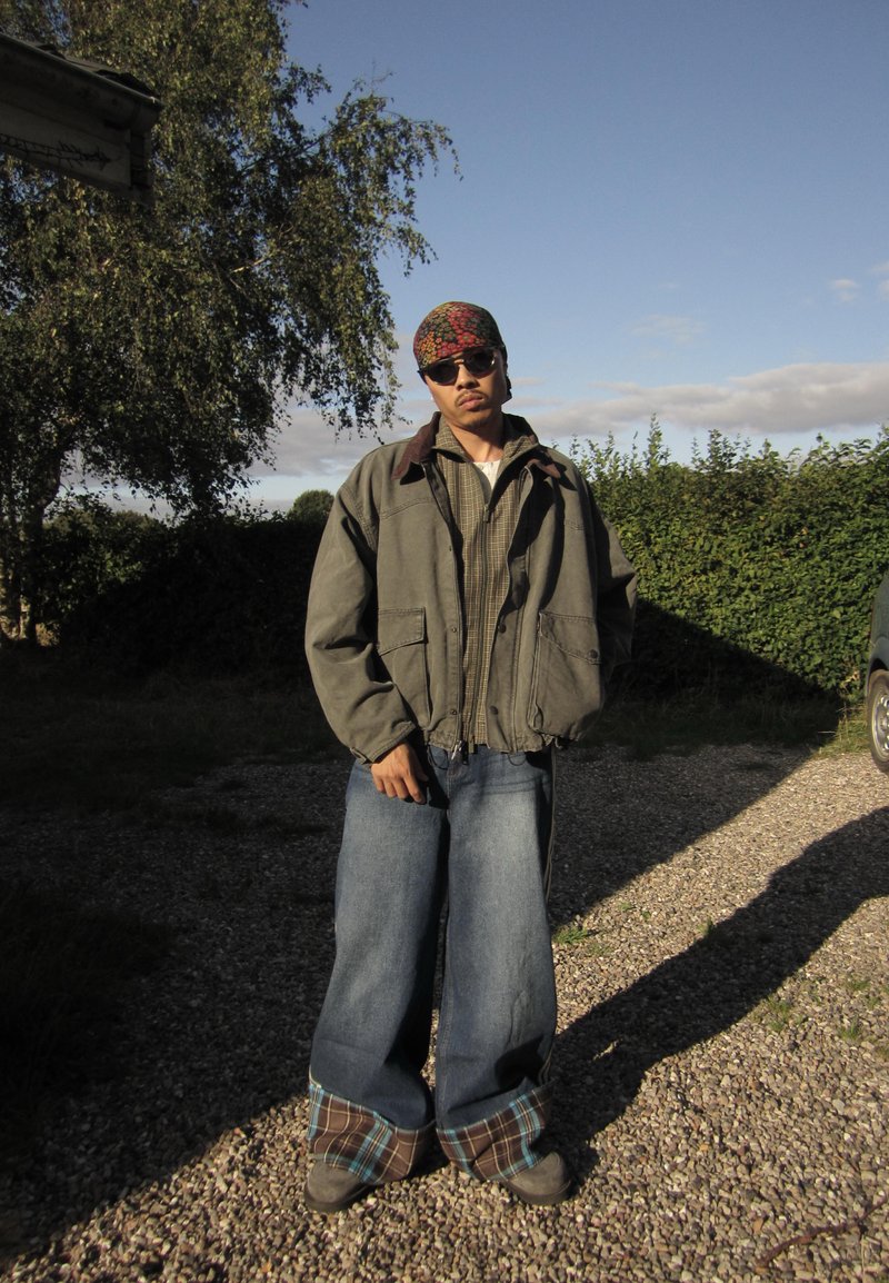 Homme portant une veste oversize, un jean ample avec des revers à carreaux, des lunettes de soleil et un foulard à motifs, debout sur un chemin de gravier avec de la verdure en arrière-plan.