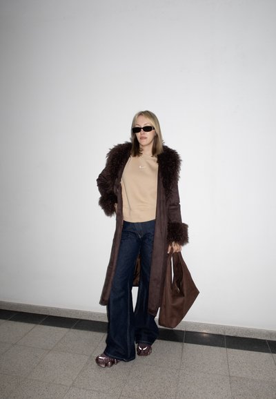 Mujer con abrigo oscuro con ribete de piel marrón, sudadera beige, vaqueros de pierna ancha, gafas de sol, sosteniendo un bolso tote marrón, de pie contra una pared blanca.