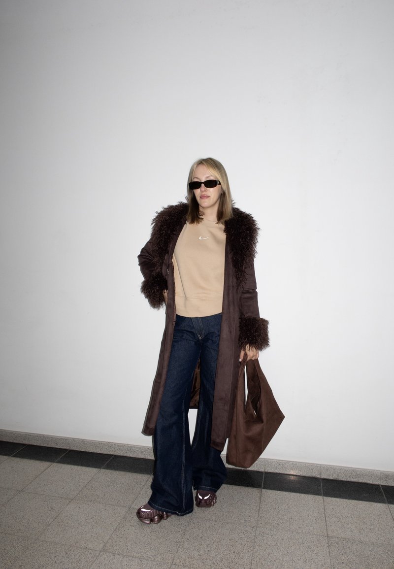 Femme portant un manteau marron foncé bordé de fourrure, un sweat-shirt beige, un jean à jambes larges, des lunettes de soleil, tenant un sac cabas marron, debout contre un mur blanc.